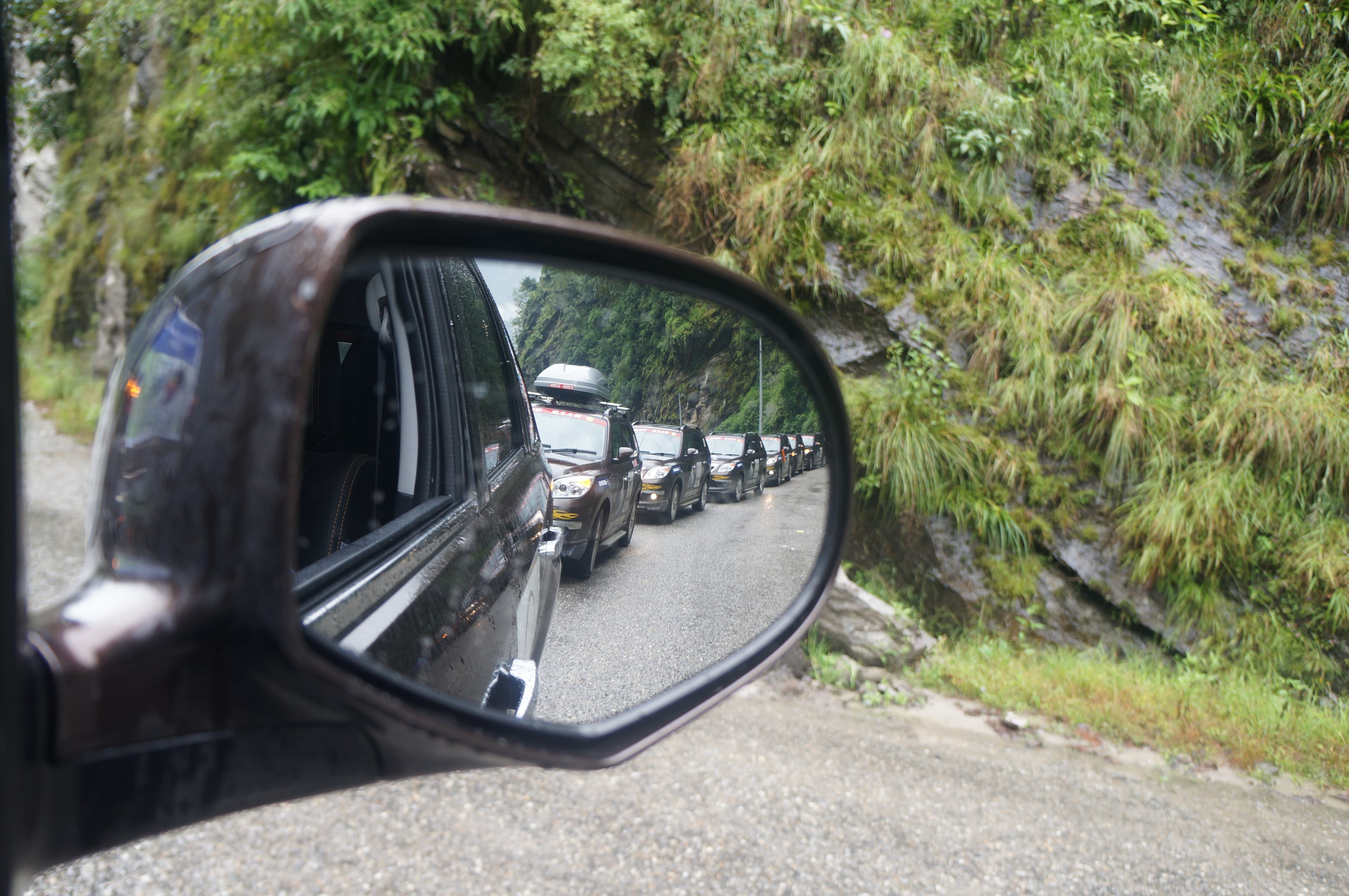 A convoy of vehicles winding along a narrow mountain road, seen in a side mirror